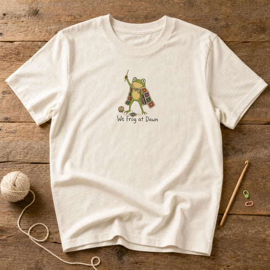 Cream We Frog at Dawn crochet frog graphic tee laid flat on a wooden table with a ball of yarn, a crochet hook and stitch markers