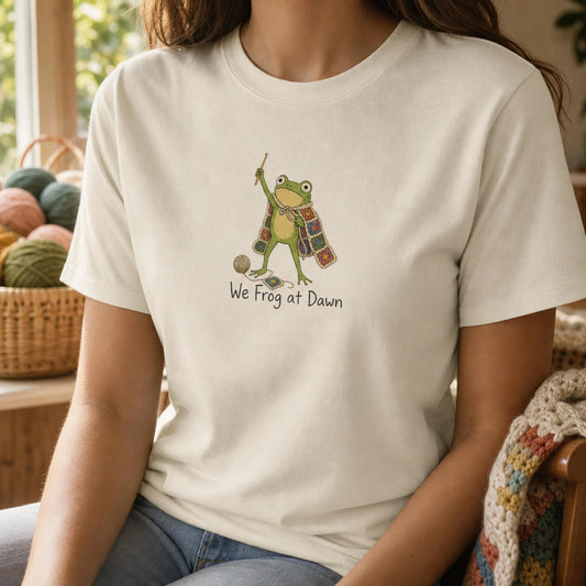 Woman wearing a cream We Frog at Dawn crochet frog graphic tee, seated beside a basket of yarn and a granny-square blanket